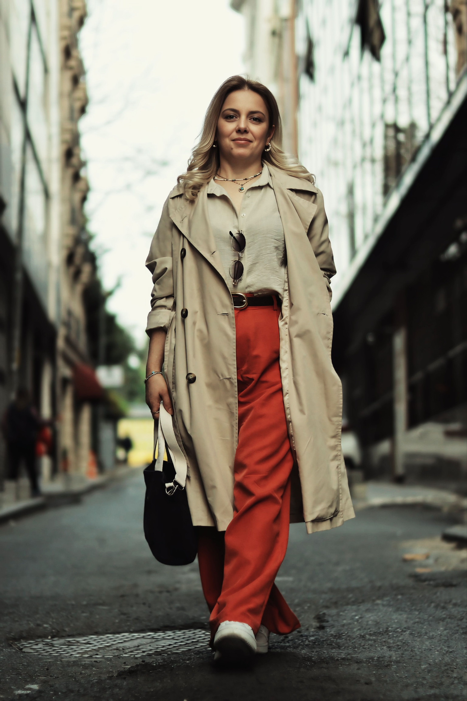A stylish woman walking on a city street in red wide-leg trousers, a trench-style jacket, layered jewelry, and carrying a black bag.